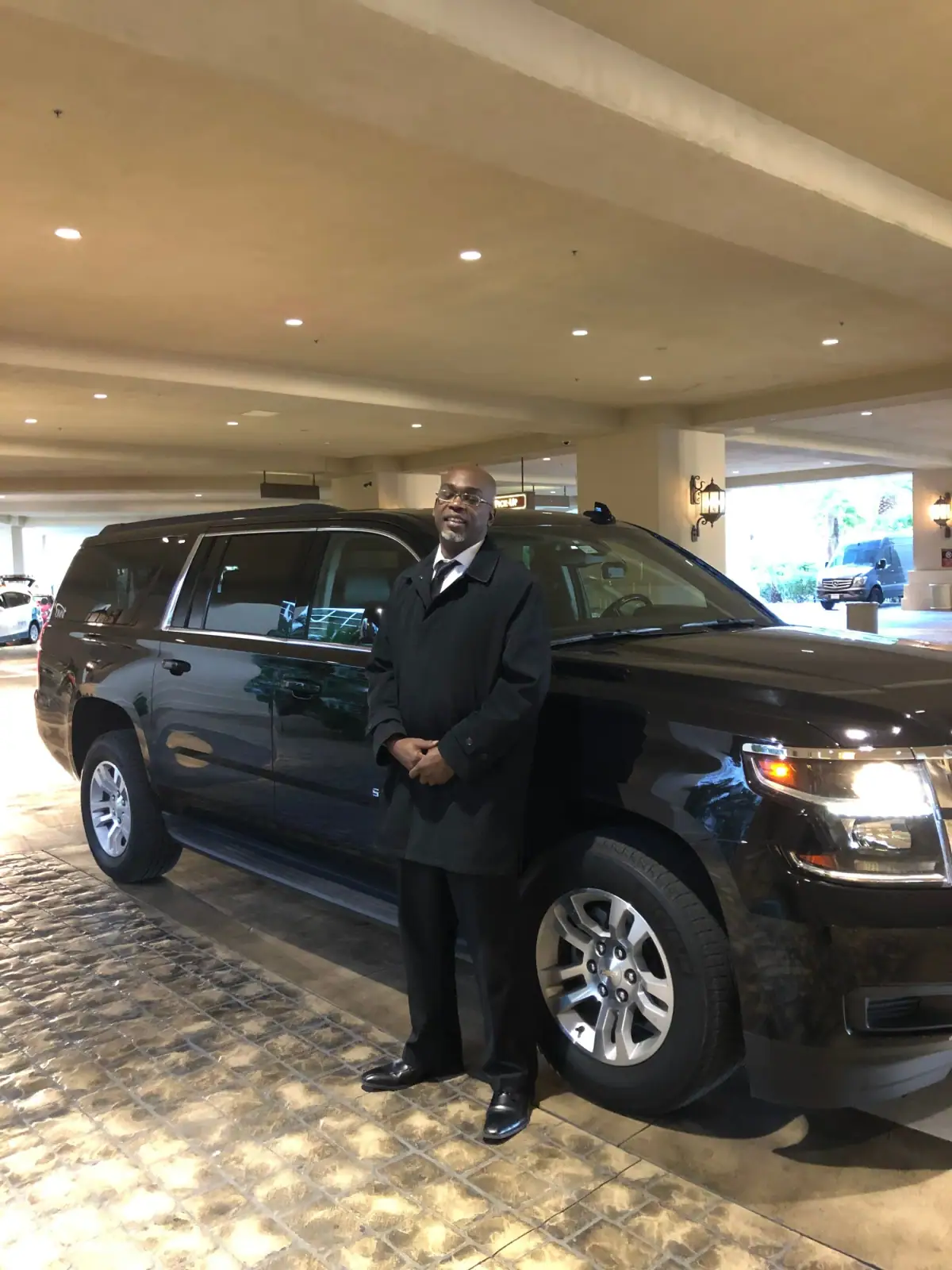 Chauffeur in a dark overcoat beside a black SUV under a hotel entrance